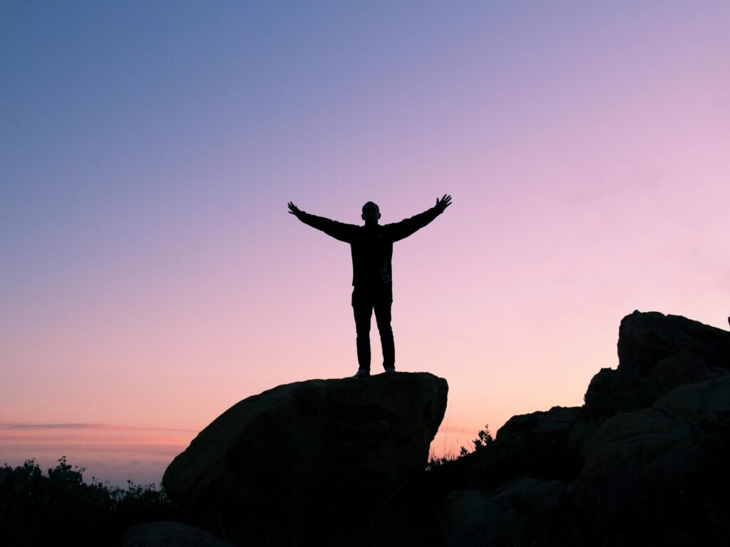 Silhouette of a man on a rock with arms raised, embracing the sunset sky.